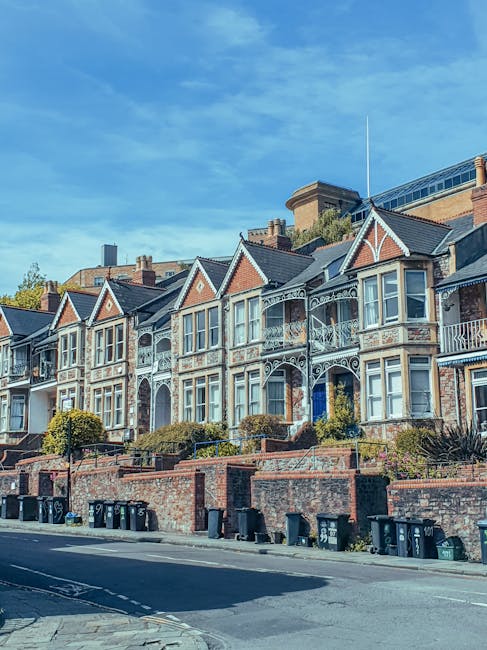 A row of Victorian-style residential houses in Ealing, featuring brick facades and decorative white wrought iron balconies. The buildings have multiple large windows, some with bay windows, and are set on a stepped hillside with small front gardens bordered by low brick walls. The street in front is paved, with several black wheelie bins lined up along the sidewalk. Bright daylight illuminates the scene, highlighting the clean and well-maintained appearance of the homes and their surroundings. Ealing Carpet Cleaning provides professional domestic and commercial cleaning services, including surface cleaning and deep cleaning to maintain hygiene and cleanliness in properties like these in the Ealing council areas.