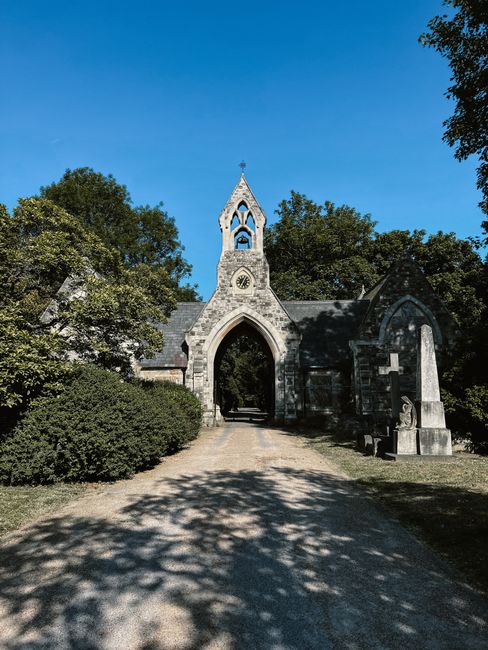 A stone church entrance with an arched gateway, topped by a small spire and decorative clock, surrounded by lush green trees and bushes under a clear blue sky. The gravel path leading to the church is partially shaded by trees, and the structure appears clean and well-maintained, reflecting careful maintenance typical of outdoor surface cleaning and preservation, as provided by Ealing Carpet Cleaning in accordance with the services highlighted on the page about avoiding hidden cleaning charges in Ealing council areas.