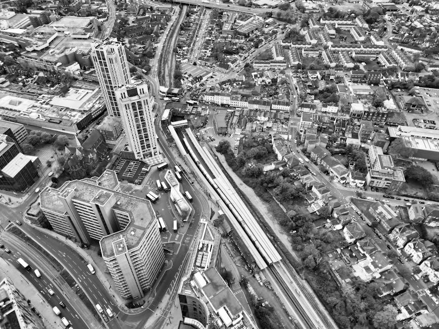 A black and white aerial view of an urban area featuring a mix of high-rise and low-rise buildings, with a busy road network including multiple lanes and vehicles. Elevated train tracks run through the center of the image, adjacent to station platforms and platforms with visible safety barriers. The surrounding streets are lined with commercial and residential structures, with some trees and open green spaces visible. Bright sunlight casts shadows, highlighting the intricate layout of the cityscape. The image captures the dense, mixed-use environment typical of central city districts, emphasizing the combination of transportation infrastructure and diverse building types. This scene could be associated with urban cleaning and maintenance, reflective of a professional cleaning service operating within a developed metropolitan context.