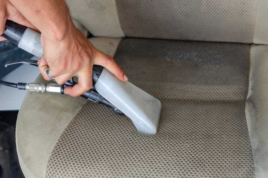 A person is performing surface cleaning on a beige, textured fabric car seat using a handheld upholstery cleaning device with a transparent cleaning head, which is emitting a focused stream of cleaning solution or steam. The device is connected by a hose to a power source, and the person's hand is steadying it while cleaning a slightly stained area of the seat. The background shows a clean, well-lit indoor environment, highlighting the process of deep cleaning and stain removal in a domestic or vehicle setting; the scene emphasizes hygiene and maintenance practices by Ealing Carpet Cleaning as part of surface sanitisation and upholstery cleaning services.