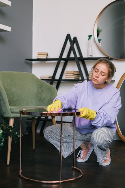 A young woman wearing a lavender sweatshirt and yellow cleaning gloves is kneeling on a dark wood floor, using a pink cloth to clean a small round glass-top side table with a metal frame, situated in a modern living room. In the background, there is a green upholstered armchair, a wooden shelf with neatly stacked books, and a large round mirror hanging on the wall, reflected in which is a plant and part of the room. The setting appears well-lit with natural light, emphasizing the cleanliness of the surface, consistent with domestic deep cleaning and surface maintenance practices by Ealing Carpet Cleaning for upholstery cleaning and stain removal tips on Pitshanger Lane.