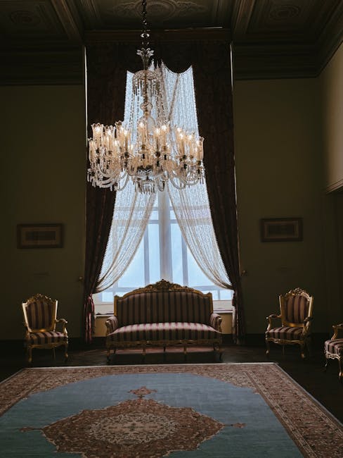 A luxurious living room with ornate dark wood paneling on the ceiling, featuring a large, elegant crystal chandelier hanging centrally. Behind the chandelier, a tall window with sheer white curtains layered with dark, heavy drapes allows natural light to filter into the space. In front of the window, a vintage-style upholstered sofa with striped fabric in muted tones is positioned, flanked by matching armchairs with similarly detailed upholstery. The room's dark wooden flooring is partially covered by a decorative area rug in shades of blue, beige, and rust, creating a classic and sophisticated ambiance. The overall aesthetic emphasizes cleanliness and maintenance, consistent with professional domestic cleaning services offered by Ealing Carpet Cleaning, as reflected in the tidy and well-preserved appearance of the furniture, drapes, and decor elements.