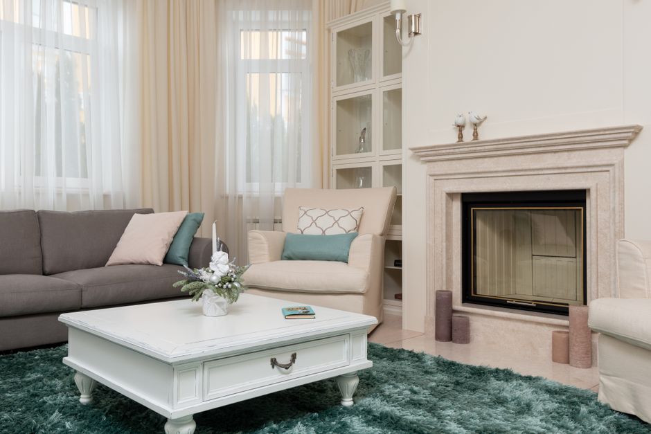 A bright and tidy living room featuring a beige armchair with a patterned cushion, a grey sofa with pastel pillows, and a white coffee table with a decorative floral arrangement and a small book on top. The room has large windows with sheer cream curtains allowing natural light to fill the space. There is a cream-colored fireplace with a simple mantel, flanked by small cylindrical decor items. To the right, a white built-in display cabinet with glass doors is partially visible. The floor is covered with a plush, dark green carpet, and the walls are painted in a neutral tone. The overall scene showcases a clean, well-maintained domestic setting, emphasizing surface cleaning and hygienic home presentation, as associated with Ealing Carpet Cleaning and their specialist cleaning services for residential properties like flats and Victorian homes.