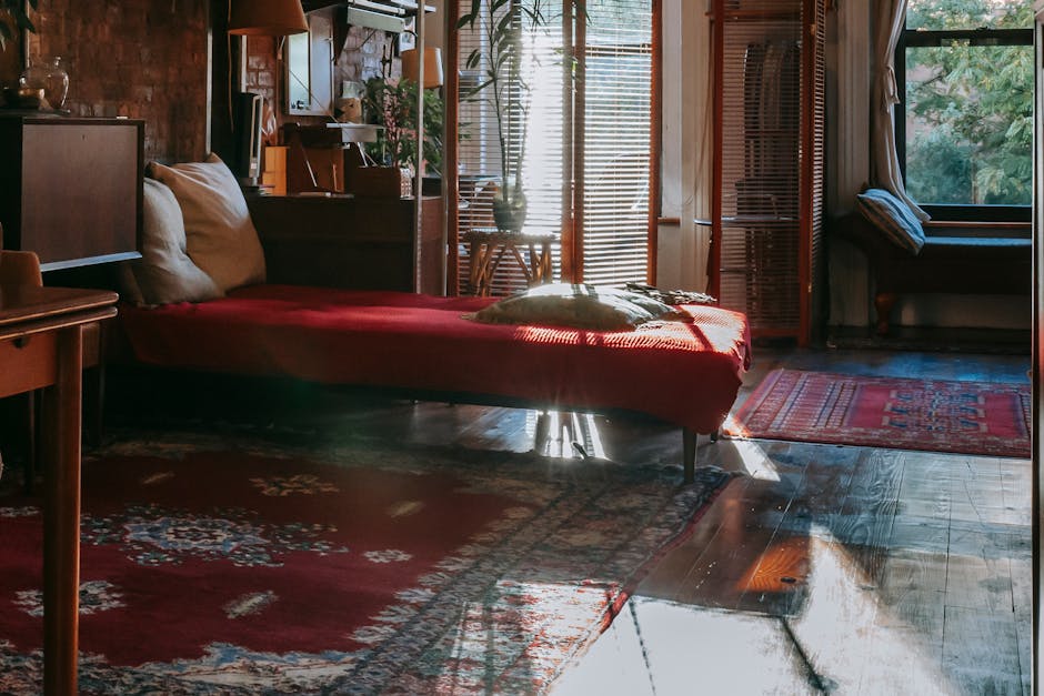 A luxurious living room with ornate dark wood paneling on the ceiling, featuring a large, elegant crystal chandelier hanging centrally. Behind the chandelier, a tall window with sheer white curtains layered with dark, heavy drapes allows natural light to filter into the space. In front of the window, a vintage-style upholstered sofa with striped fabric in muted tones is positioned, flanked by matching armchairs with similarly detailed upholstery. The room's dark wooden flooring is partially covered by a decorative area rug in shades of blue, beige, and rust, creating a classic and sophisticated ambiance. The overall aesthetic emphasizes cleanliness and maintenance, consistent with professional domestic cleaning services offered by Ealing Carpet Cleaning, as reflected in the tidy and well-preserved appearance of the furniture, drapes, and decor elements.