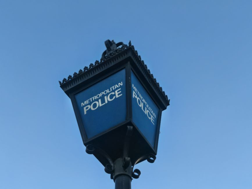 A close-up view of a traditional Victorian-style street lamp with a black metal pole and ornate decorative elements. The lamp housing is rectangular with blue panels displaying the white text 'METROPOLITAN POLICE' on two visible sides. The lamp is mounted high on the pole against a clear blue sky, indicating outdoor urban surroundings. The surface of the lamp appears well-maintained and clean, with no visible graffiti or damage, emphasizing its functional and aesthetic condition as part of the city street’s infrastructure. The lighting suggests it is daytime with natural sunlight illuminating the lamp evenly. This image is associated with Ealing Carpet Cleaning's services, highlighting a typical street scene that may relate to access challenges in urban cleaning jobs.