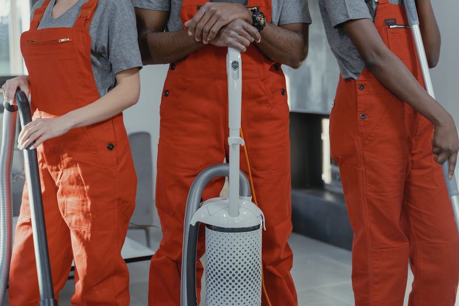 Three professional cleaners from Ealing Carpet Cleaning are shown in an indoor setting, dressed in grey shirts and bright orange uniforms, each holding a vacuum cleaner with grey hoses. The image focuses on their lower torsos and hands, with no visible faces. They are positioned in a well-lit room with a modern background, featuring tiled flooring and a dark counter or furniture piece behind them. The surfaces appear clean and well-maintained, and the vacuums are ready for surface cleaning or deep cleaning tasks. The image emphasizes the hygiene and maintenance aspects associated with domestic cleaning services, highlighting tools used by professional cleaners in a typical residential environment.